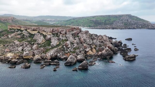 jagged rock stacks dot the shallows off a reddish headland on maltas coast sea caves undercutting the cliff face above a sandy cove rolling green farmland under grey overcast sky