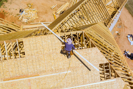 Worker stands on roof uses tools to attach materials while construction continues on new house nearby.