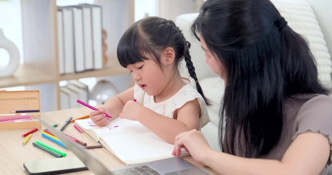 Asian mom points across notebook while preschool daughter draws with pink pencil beside laptop and crayon, building fine motor control, child communication, confidence, and education connection.
