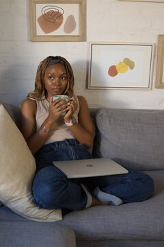African American woman sitting on gray couch at home holding ceramic mug with laptop on lap