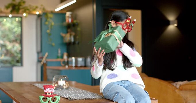 Putting on Merry Christmas glasses, Indian girl shaking gift box at home to hear surprise inside