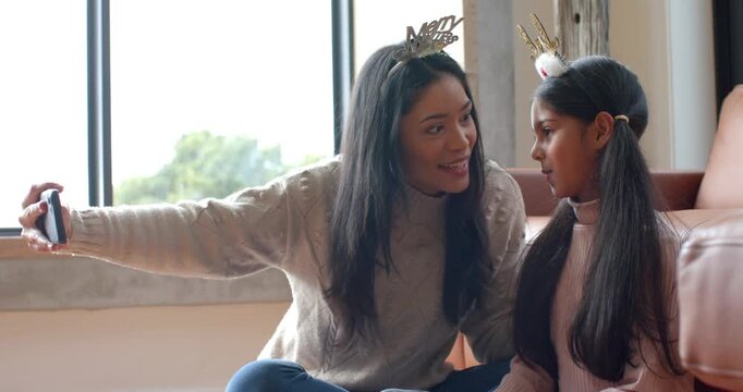 Mother wearing tiara holding phone guiding daughter leaning selfie at home beside gift box laughing