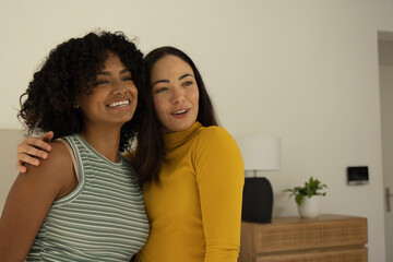 Obraz premium Diverse female friends standing arm-in-arm at home wearing striped and mustard tops beside dresser