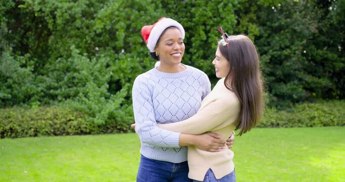 Diverse female friends spotting Santa hat, festive headband, hugging in garden, pointing toward sky