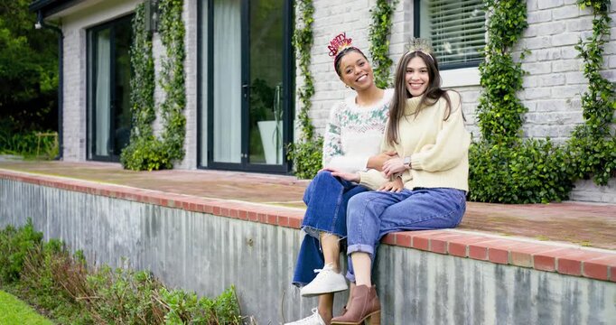 Camera zooming in Diverse female friends fixing crowns linking hands on brick ledge for Christmas