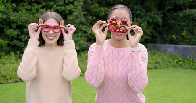 Female friends adjusting red deer ear and Christmas tree glasses in garden celebrating holiday fun
