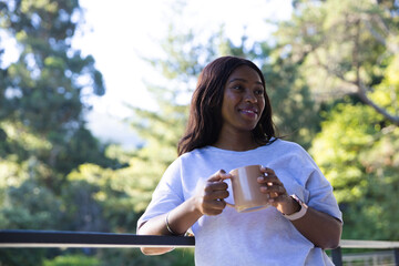 African adult woman cradling mug leaning on metal rail on balcony, wearing smartwatch, bracelet © WavebreakMediaMicro