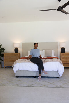 African woman sitting on bed in minimalist bedroom wearing blue tee black leggings pink sneakers