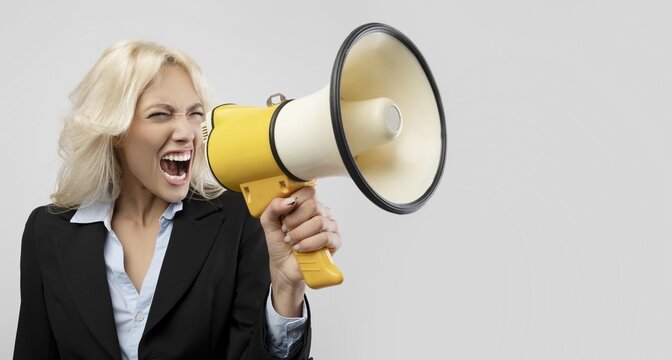 Angry Caucasian Woman In Business Suit Yelling Into Megaphone At Office, Displaying Strong Emotion While Motivating Colleagues In A Professional Setting, Business Concept.