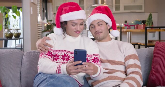 African American couple sitting on sofa in Santa hats, reacting, laughing at clip on blue phone