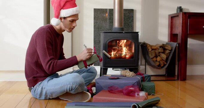 African American man putting on red Santa hat, labeling green gift for holiday beside wood stove
