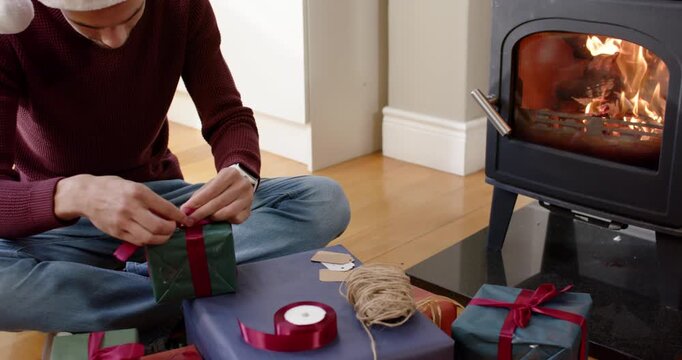 Man sitting by woodstove tying red ribbon on green box, donning Santa hat, preparing gifts
