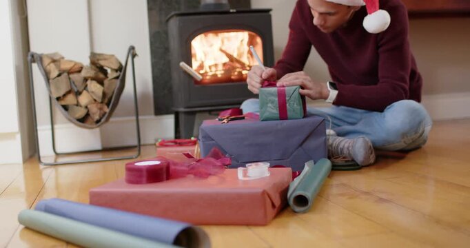 Camera revealing African-American man in Santa hat wrapping gifts on floor by stove for gifting