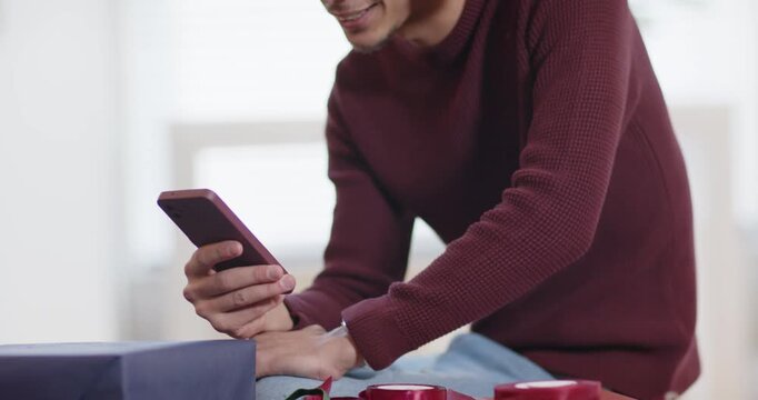 Man at table holding phone, seeing message, donning Santa hat and arranging gifts for holiday