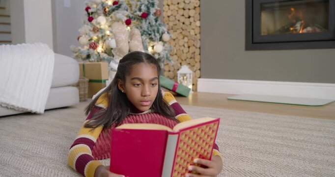 African American teen lying on rug reading red book, finding funny line by Christmas tree, laughing
