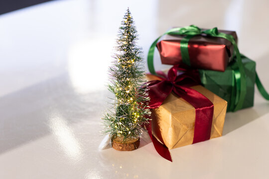 Miniature tinsel Christmas tree with warm lights standing on tabletop next to metallic gift boxes