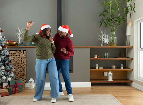 African American couple dancing by fireplace at home wearing Santa hats near Christmas tree