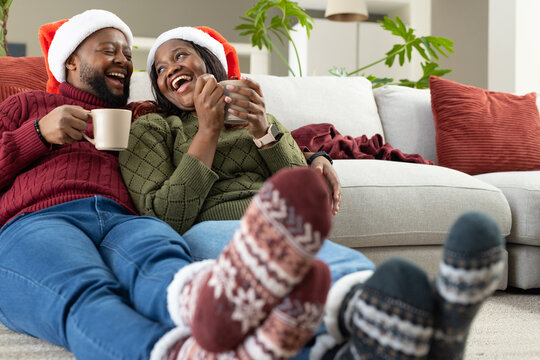 African American couple laughing sipping mugs wearing Santa hats and patterned socks in living room