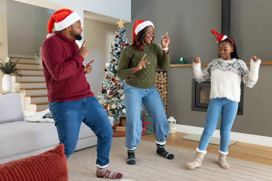 Family of three with youth dancing by Christmas tree at home wearing sweaters and Santa hats