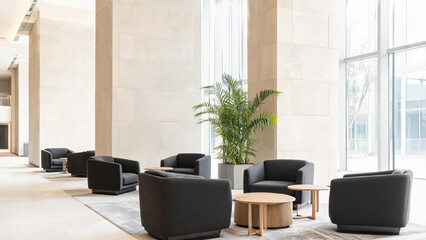 Fototapeta premium A bright modern indoor lobby seating area with dark armchairs, small tables, a large potted plant, and light stone walls and floors.