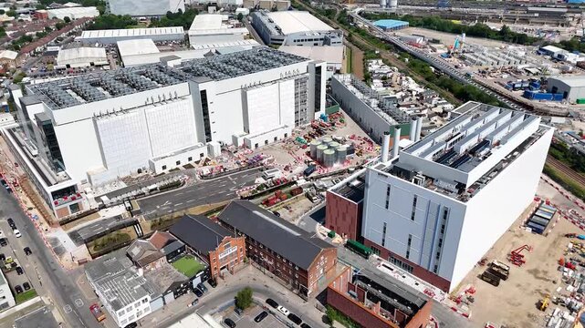 Two data centre buildings with rooftop chiller plant stand behind rows of terraced houses in west London, with construction works and lorry yards alongside