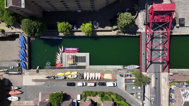 Close aerial view of a red bascule bridge over a green dock channel, with kayaks and dinghies stacked on the quay below