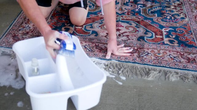 Woman scrubbing a decorative rug with a soapy brush on concrete floor.