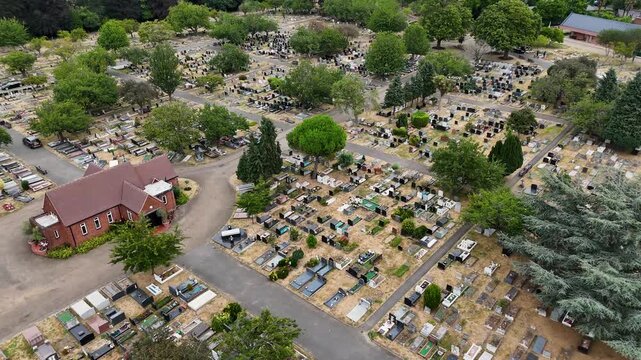 Small chapel stands among rows of graves and mature trees in a large cemetery with parched grass between the plots