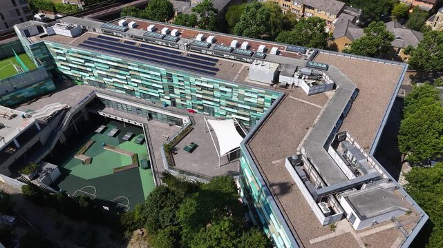 Modern school building wraps around a green multi use sports court, with solar panels and a canopy on its roof terraces