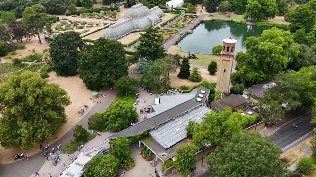Victorian glasshouse and a tall brick campanile stand beside a lake in botanic gardens, with the lawns burnt brown