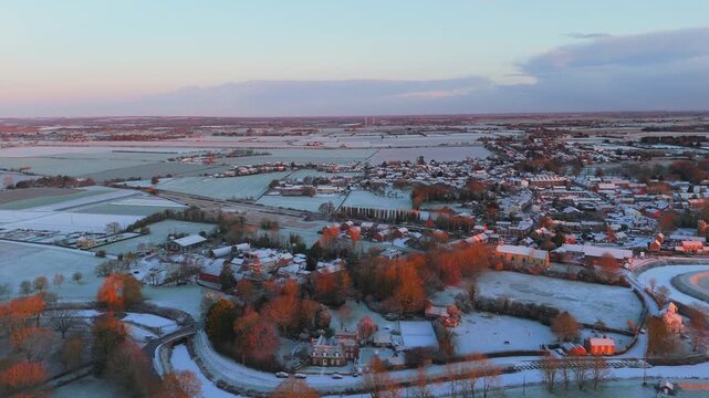 Aerial shot shows snow-blanketed village at sunrise. Autumn trees with orange foliage stand out among homes. Frozen fields stretch into distant countryside. Perfect for holiday, travel