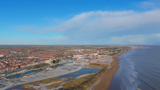 Clear blue sky with wispy clouds above the sea. Sandy beach meets gentle waves along the coastline. Red-tiled town sprawls towards the horizon. Urban area blends with natural coastal features