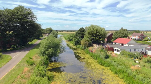Aerial view above calm canal with green algae floating on surface. Lush trees and grasses border the waterway. Small homes with red roofs dot the countryside. Perfect for nature, rural