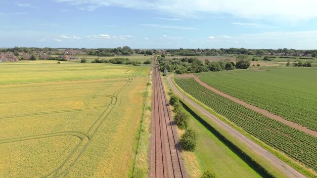 Aerial view of rural tracks between golden and green fields. Clear blue sky above tranquil countryside landscape. Sunlit fields stretch wide, bordered by railway lines. No people, just quiet