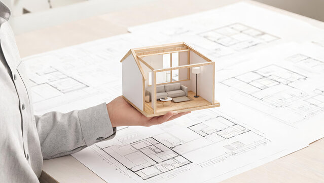 A professional holds a miniature house model above architectural floor plans, illustrating residential design and planning work.