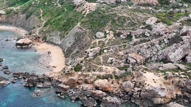 sandy cove beach hikers walking shoreline switchback hiking trail scrubland hillside rocky headland malta west coast secluded bay aerial drone footage coastal hiking tourism