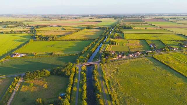 Golden light paints vast green fields stretching to the horizon. Winding canal cuts through the countryside, flanked by trees. Bridge crosses the water, connecting distant farmland. Sibsey Trader