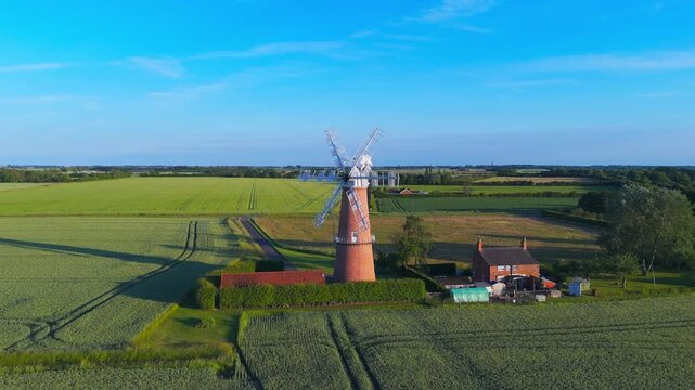 Ancient windmill stands tall amid green fields. Blue sky stretches over rural countryside. Sunlight casts long shadows on the land. Windmill's red brick contrasts with green crops.