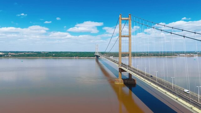 Massive suspension bridge spans wide river under clear sky. Bridge towers rise high, cables stretch taut across water. Humber suspension bridge over the river in Humberside, UK