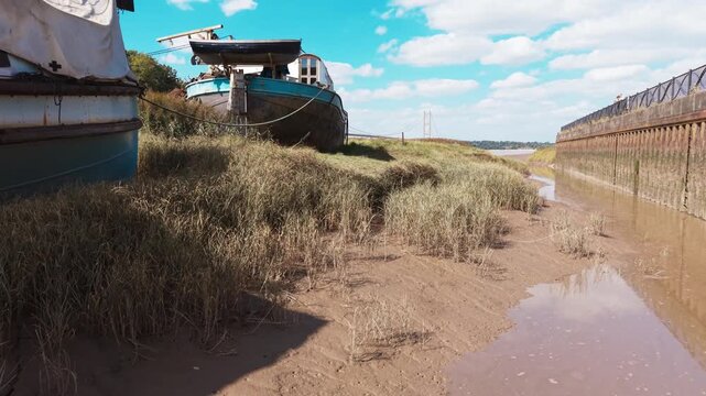 Sunlit sandbank holds weathered boats, rusted and quiet. Tall grasses sway gently, framing muddy waterways. Distant wall stretches. Humber suspension bridge over the river in Humberside, UK