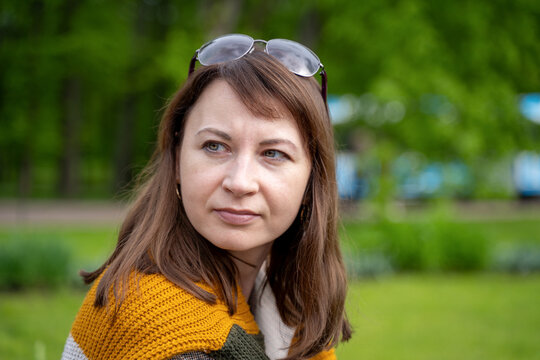 An adult woman with brown hair and sunglasses pushed up on her head wears a mustard knitted shawl while looking sideways in a park.