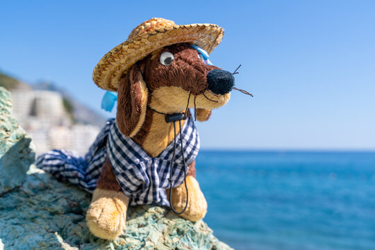 A brown plush dachshund toy dressed in a checkered shirt and straw hat with googly eyes and a twig mustache perches on a rock above the sea, a playful travel adventure mascot.