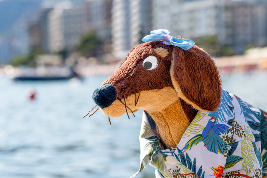 A brown plush dachshund toy with googly eyes, a blue hair bow and a tropical print shirt poses against a sparkling bay with a boat and high rise skyline behind it, a whimsical mascot for coastal city.