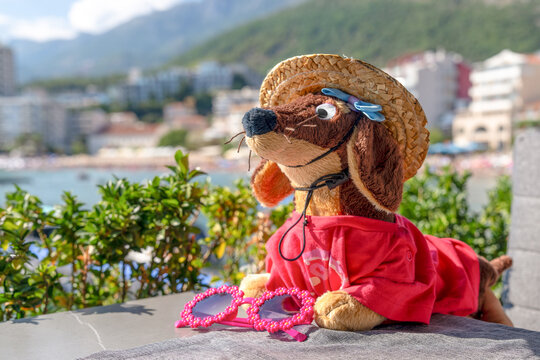 A stuffed dachshund toy wearing a straw hat and red shirt lies on a ledge beside pink heart shaped sunglasses, with a seaside town and mountain visible beyond.