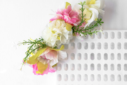 A decorative garland of artificial pink, white and yellow flowers with green foliage lies diagonally on a white perforated surface, a concept of handmade floral craft.