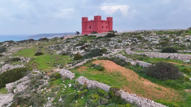 bright red crenellated watchtower hilltop fortress drystone walled terraces stone corbelled hut malta countryside aerial drone footage coastal fortification heritage