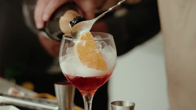 Bartender adding an orange slice and cherry to a red cocktail over ice in a stemmed glass