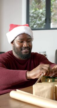 Vertical video: Santa hat urging African American man tying ribbon and tagging for gifting at table
