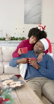 Vertical video: Diverse couple on sofa raising red mugs for toast after woman entering with antlers