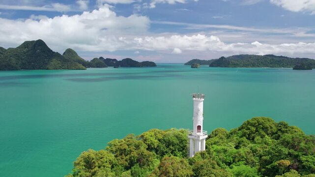 Cinematic aerial view of a remote lighthouse rising above tropical forest with brilliant turquoise waters and multiple lush islands extending across the Malaysian seascape.